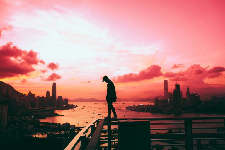 Sylwetka człowieka stojącego na barierce z widokiem na miasto i zatokę podczas zachodu słońca z różowym niebem. Czatboty AI sprawiły, że jest mniej samotny