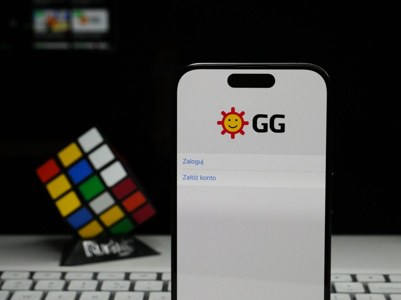 Smartfon z otwartą aplikacją o nazwie "GG" oraz ikoną uśmiechniętego słońca na ekranie, w tle niewyraźny kostka Rubika i klawiatura komputera.