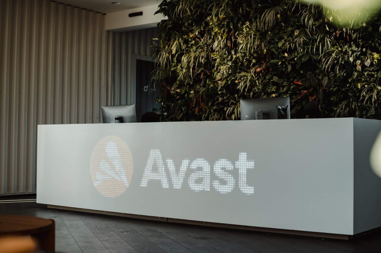 Recepcja z logo firmy Avast podświetlone na białym blacie, z roślinnością w tle oraz komputerami na blacie recepcji.