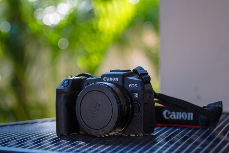 Aparat fotograficzny marki Canon model EOS R położony na metalowej siatce z rozmytym tłem zieleni.