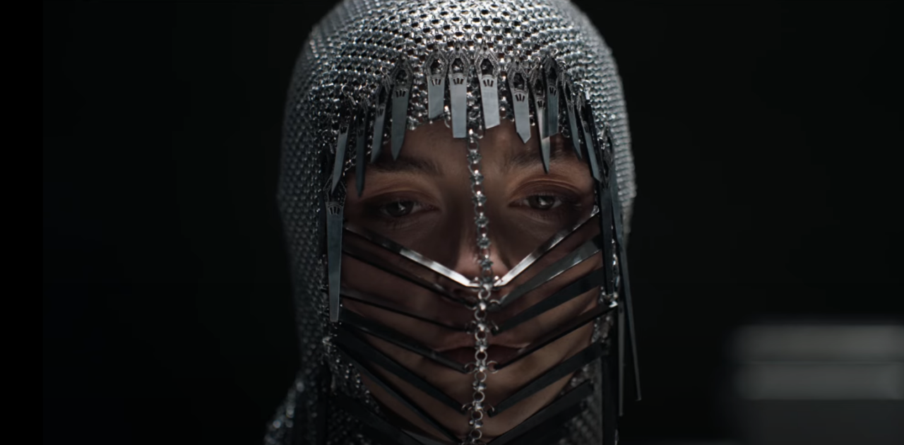 Osoba nosząca metalową zasłonę na twarz z długimi, cienkimi elementami przypominającymi kolce oraz kaptur z kryształkami na ciemnym tle.