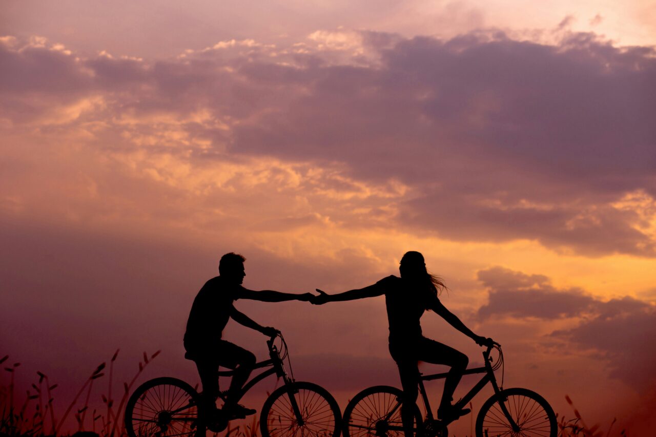 Sylwetki dwóch osób na rowerach trzymających się za ręce na tle zachodzącego słońca i pomarańczowo-różowego nieba. Oszustwa romantyczne nie są im znane