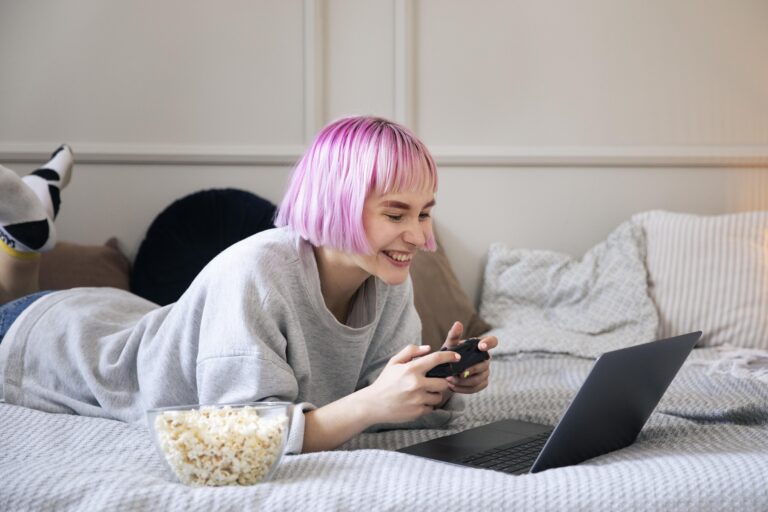 Osoba z różowymi włosami używa smartfona leżąc na łóżku obok laptopa i miski z popcornem, tak spędza Walentynki.