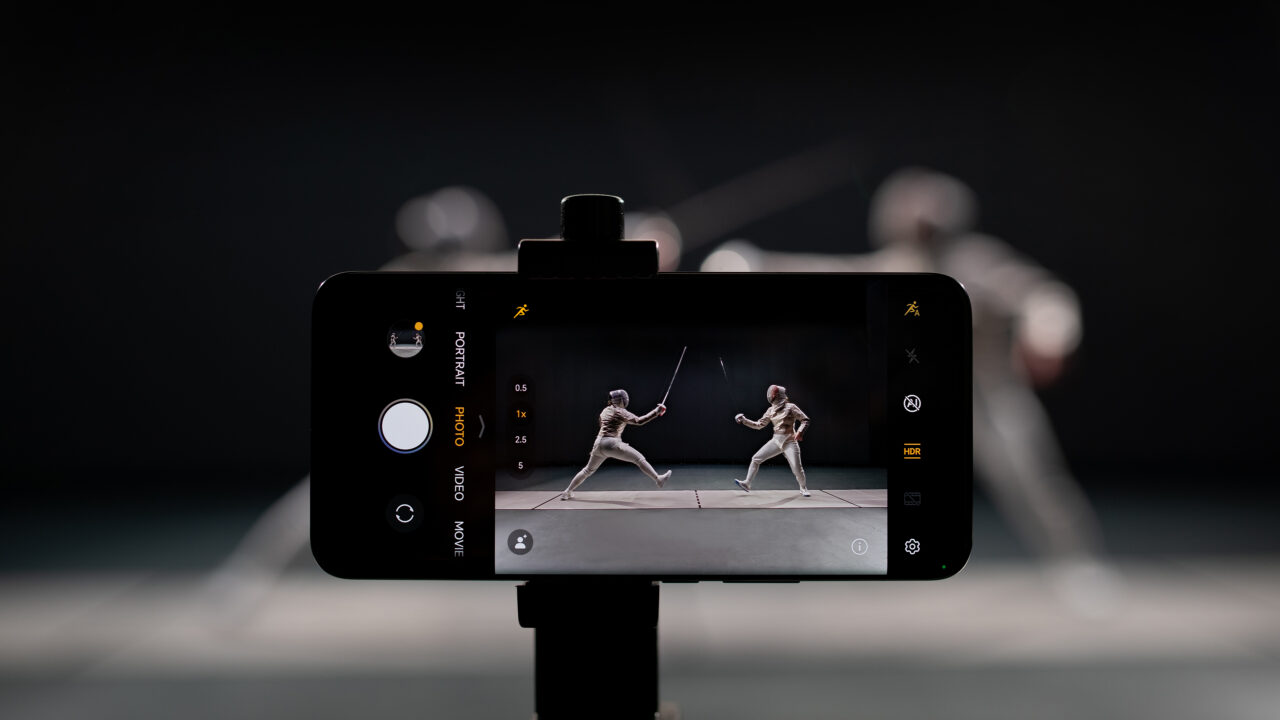 Smartfon umieszczony na statywie robi zdjęcie dwóm małym figurkom szermierzy, ustawione są one na tle rozmytego ciemnego wnętrza.