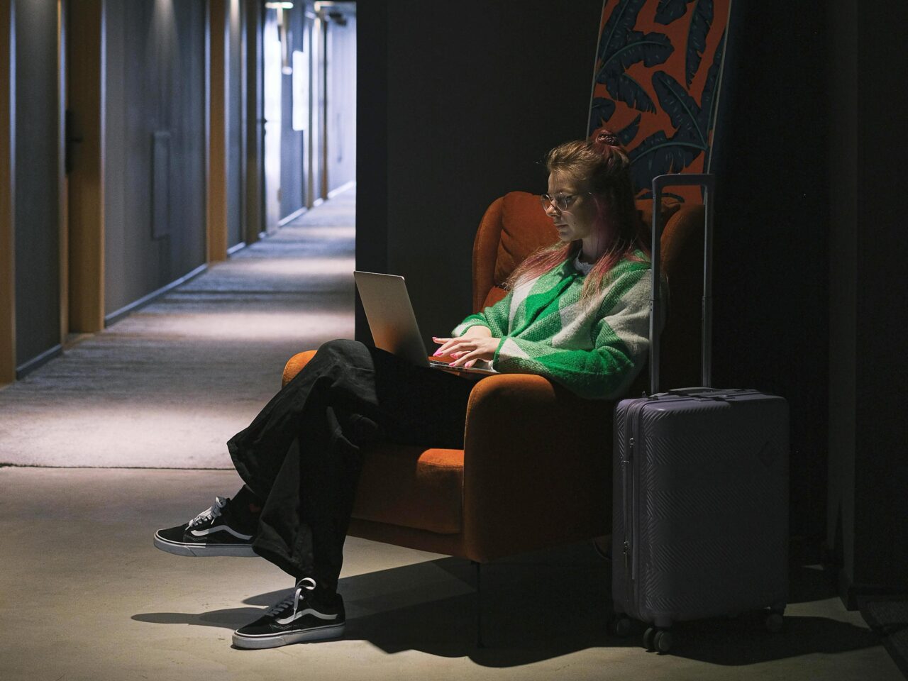 Kobieta w czerwonym fotelu pracuje na laptopie w ciemnym hotelowym korytarzu, obok niej stoi walizka na kółkach.