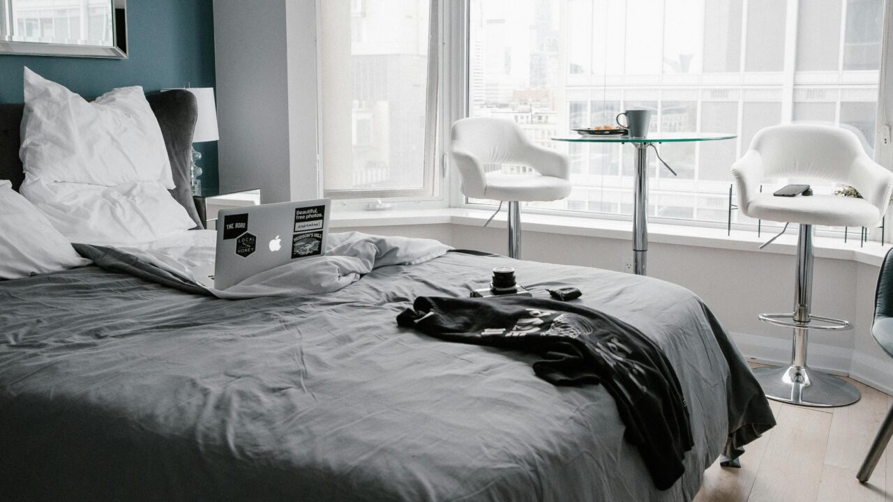 Pokój hotelowy z łóżkiem, na którym leży otwarty laptop i czarna koszulka, z widokiem na okno i dwoma białymi krzesłami przy stoliku.