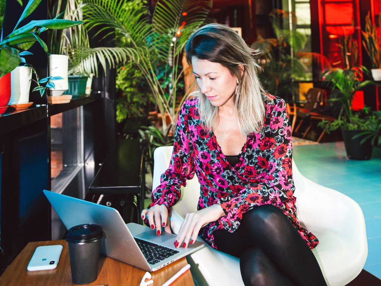 Kobieta pracująca na laptopie w wnętrzu z roślinami, obok niej kubek do kawy i telefon komórkowy.