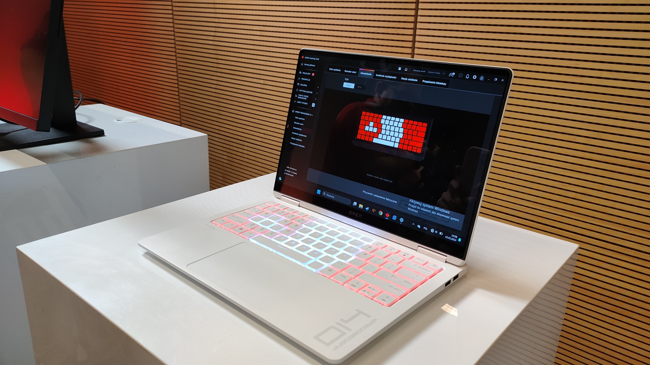 Laptop gamingowy HP Omen Transcend 14 na białym blacie z podświetlaną klawiaturą w czerwonych i białych barwach, z otwartym oprogramowaniem do dostosowywania ustawień klawiatury na ekranie, znajdujący się na wystawie z drewnianym tłem w pionowe linie.