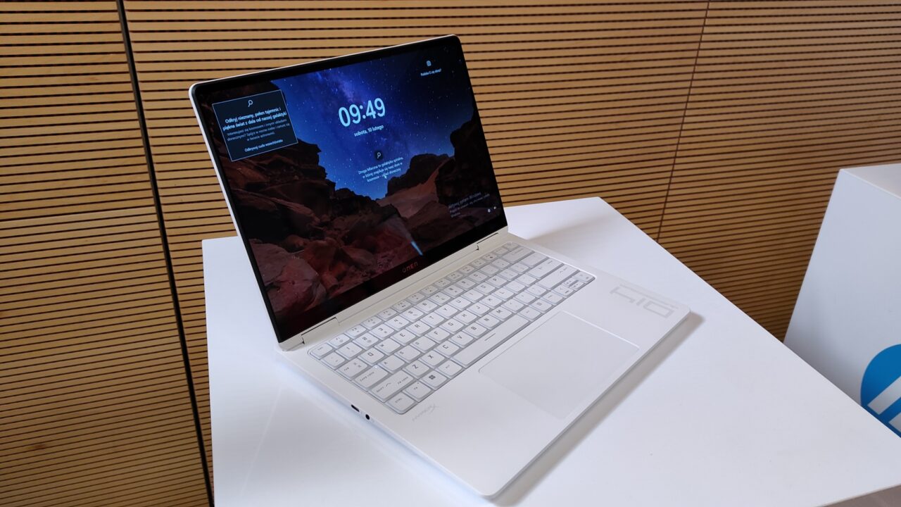 Laptop HP Omen Transcend 14 z białą klawiaturą i ekranem wyświetlającym ekran blokady z grafiką kosmiczną, oparty o tekturową podstawkę na biurku z jasnobrązowym tłem.