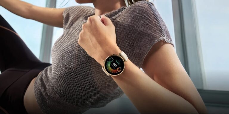 Kobieta mająca na nadgarstku smartwatch z kolorowym ekranem pokazującym dane fitness, trzyma się drugą ręką za ramię w pomieszczeniu z naturalnym światłem.