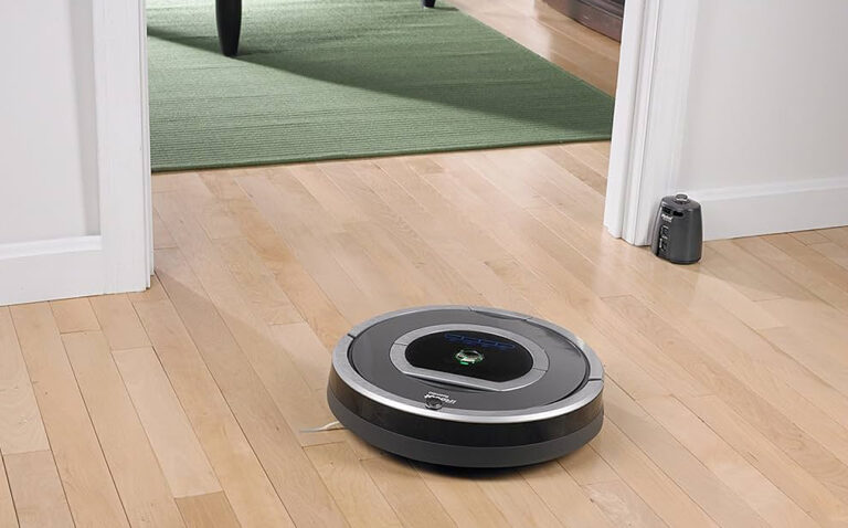 Robot odkurzający iRobot Roomba pracujący na drewnianej podłodze w domowym wnętrzu, w pobliżu progu przejścia na dywan.