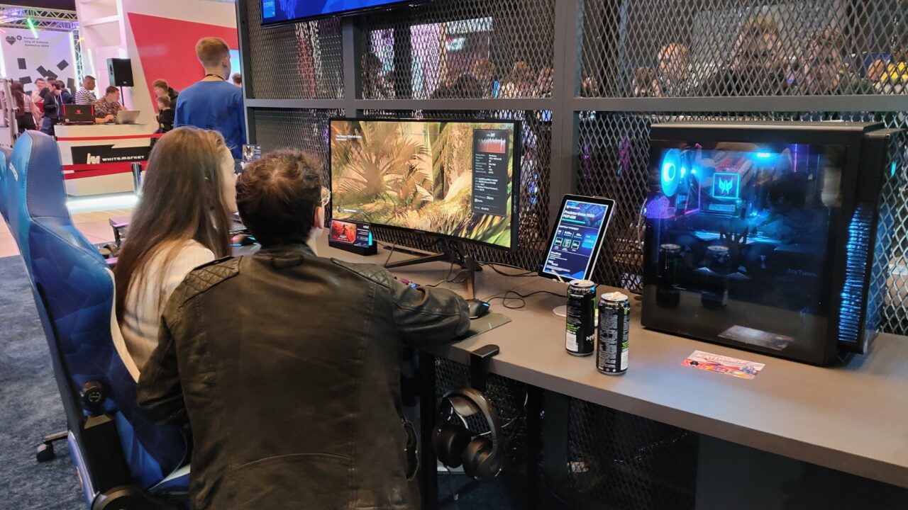 Dwie osoby grające w grę komputerową na targach technologicznych, otoczone przez różnorodne sprzęty do gier i promocyjne materiały.