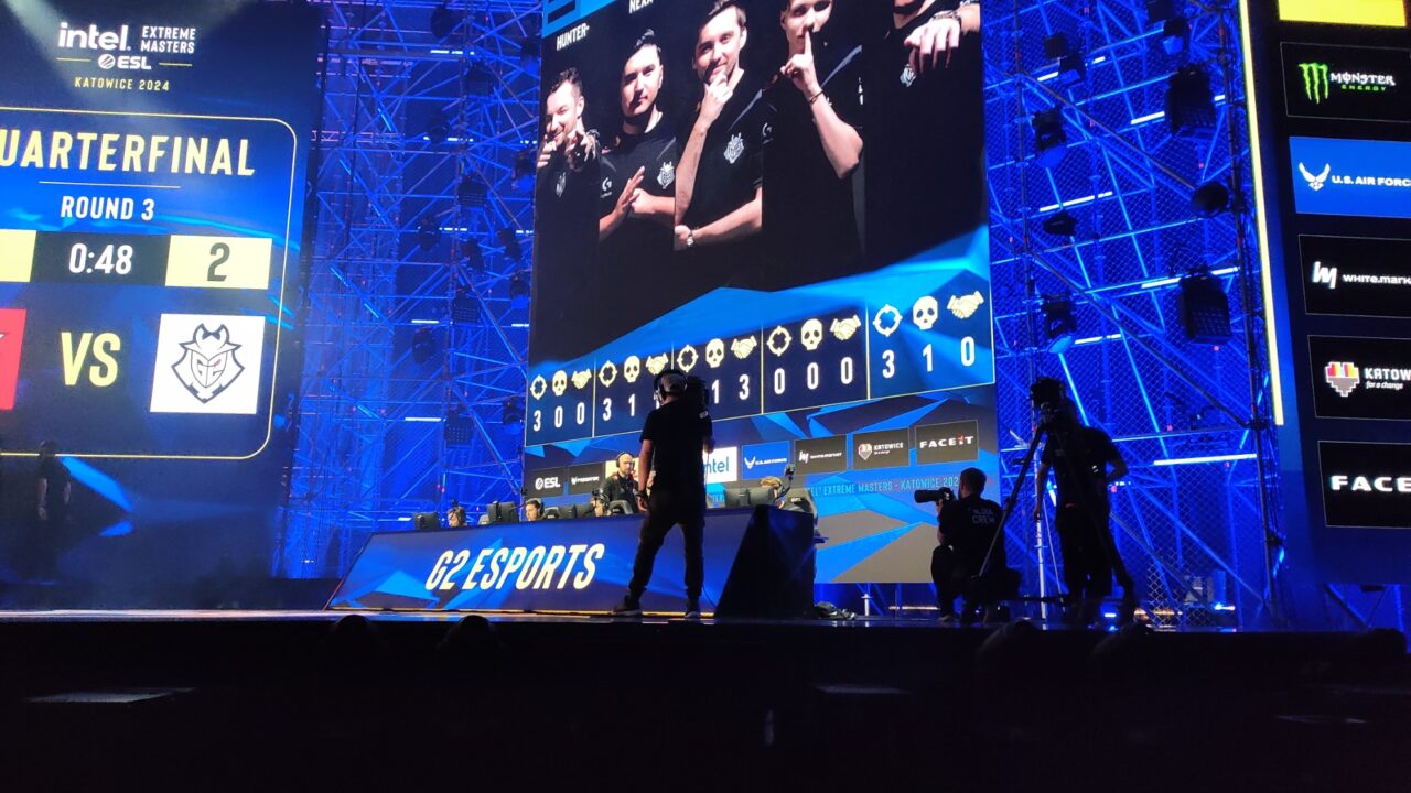 Scena e-sportowego wydarzenia IEM Katowice 2024 z dużym ekranem wyświetlającym wyniki meczu pomiędzy dwoma drużynami, a na pierwszym planie widać sylwetki osób fotografujących wydarzenie.