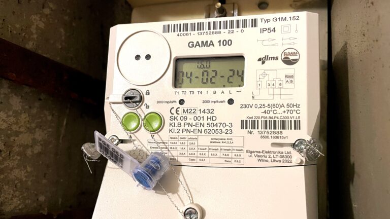 Licznik elektryczności GAMA 100 zamontowany na ścianie z wyświetlaczem pokazującym aktualny czas i datę oraz różne parametry techniczne.
