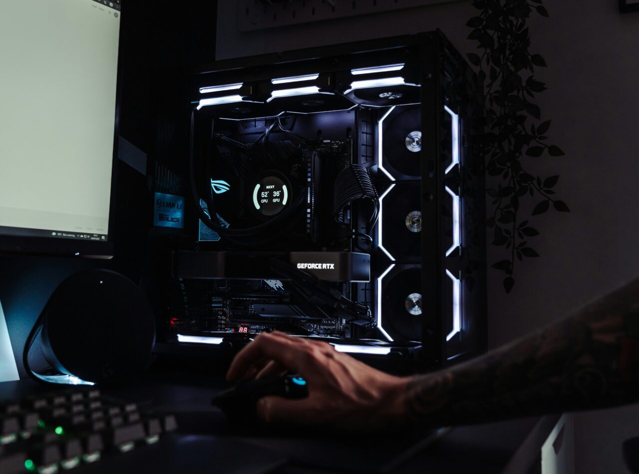 Komputer gamingowy z oświetleniem LED, obok osoba używająca myszki, w tle monitor i czarna klawiatura.