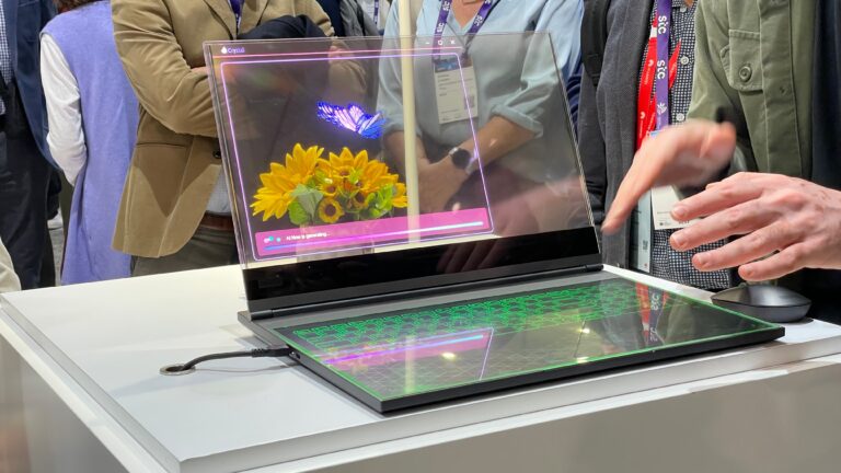 Laptop z przezroczystym ekranem OLED prezentowany na targach, na którym wyświetlany jest obraz słoneczników, w tle widoczni ludzie oraz ręka osoby manipulującej urządzeniem.