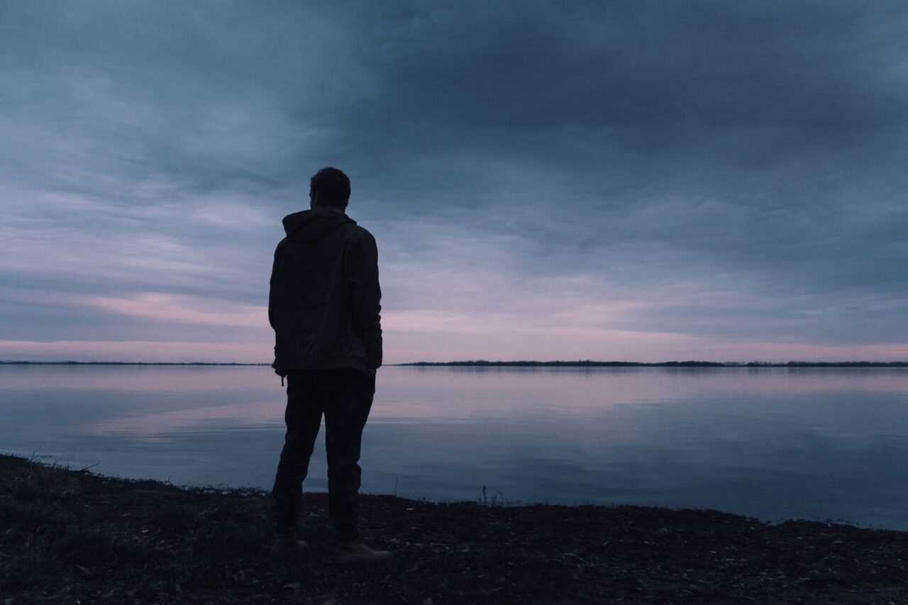 Mężczyzna stojący na brzegu jeziora podczas zmierzchu z widokiem na spokojną wodę i niebo w odcieniach błękitu i różu.