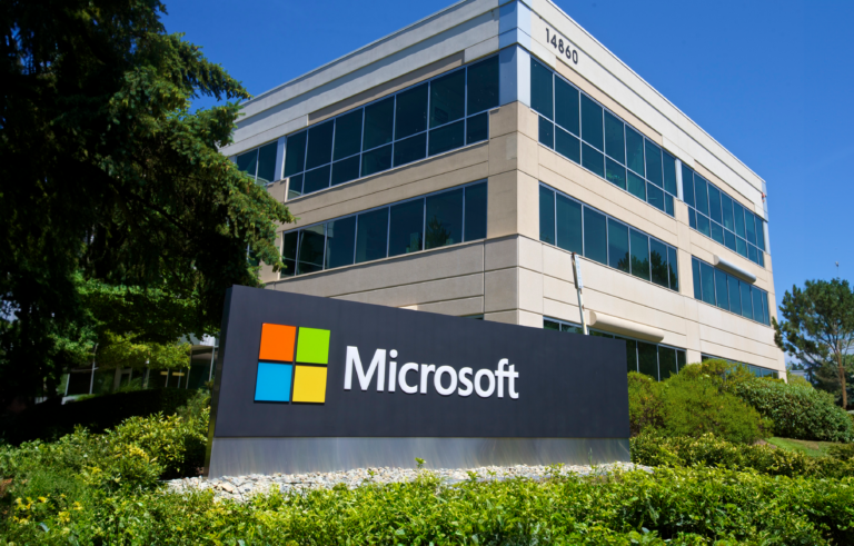 Tablica z logo firmy Microsoft przed budynkiem biurowym otoczonym zielenią.