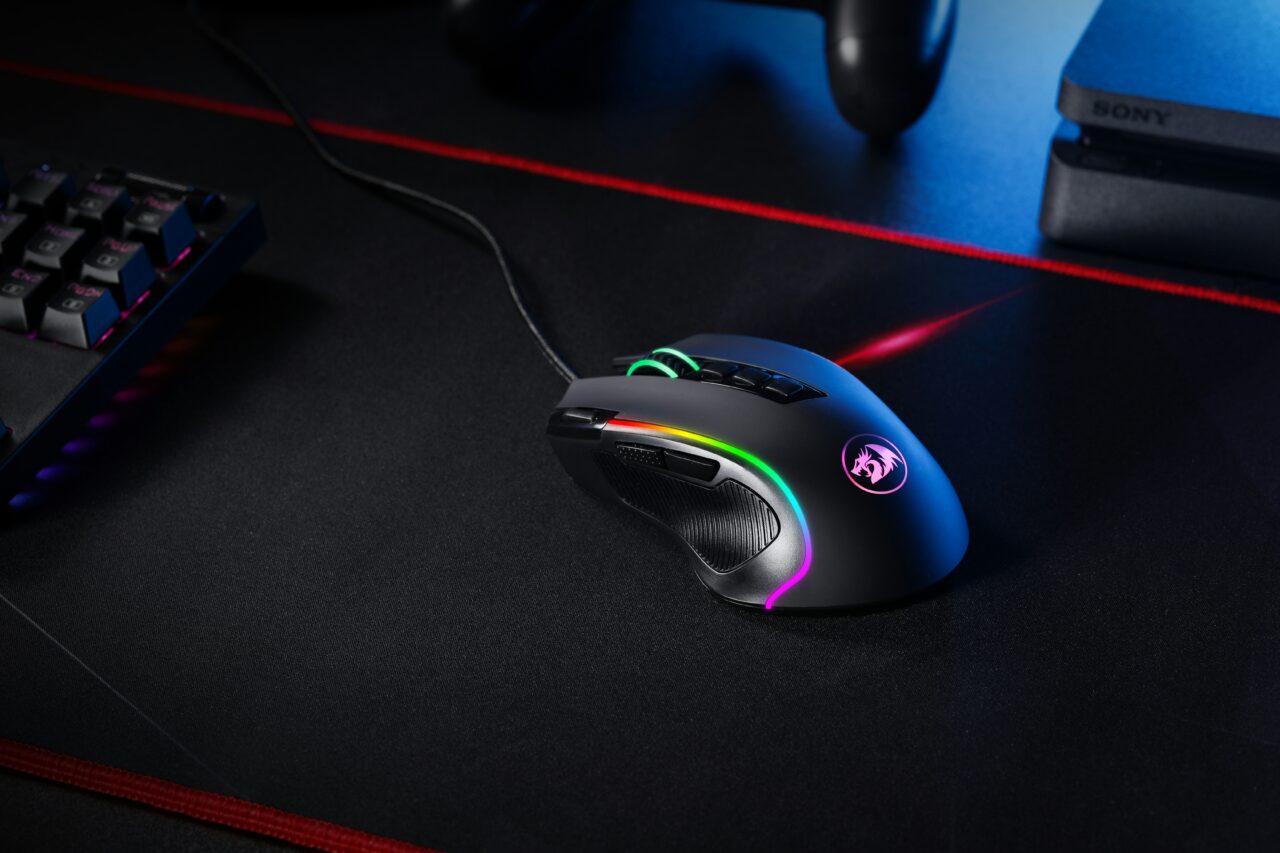 Gamingowa mysz komputerowa z podświetleniem RGB na ciemnym biurku, obok klawiatura i konsola do gier.