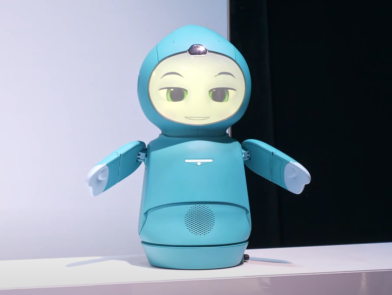 Robot o wyglądzie antropomorficznym z dużymi okrągłymi oczami i w niebieskim kolorze, stojący na białym blacie z ciemnym tłem.