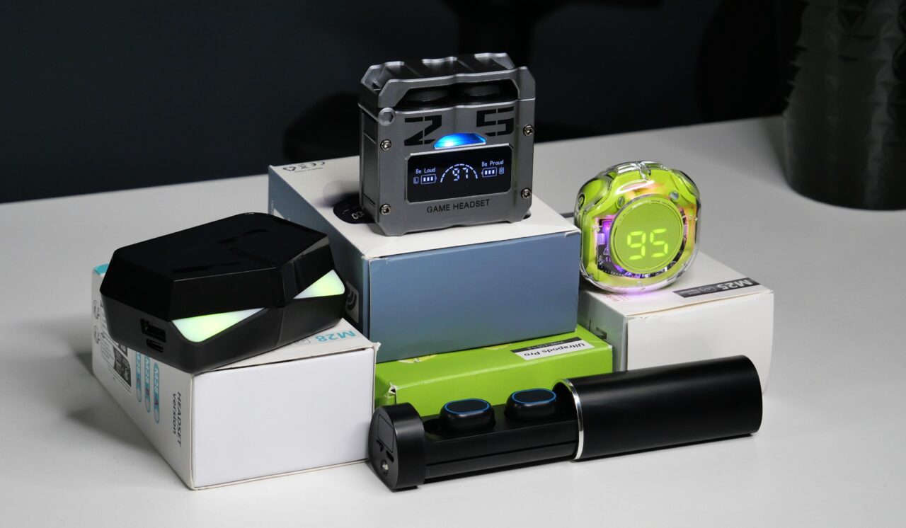 Zbiór nowoczesnych gadżetów elektronicznych, w tym gogle VR, zegar z elektronicznym wyświetlaczem i budzik w stylu militarystycznym, położone na kolorowych pudełkach na białym stole.