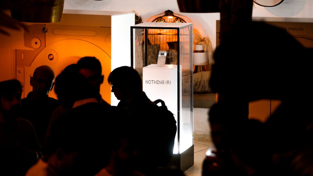 Sylwetki ludzi w półmroku z przodu, z wystawą produktu w przezroczystej witrynie ze smartfonem Nothing Phone (2a) świecącej na tle pomieszczenia.