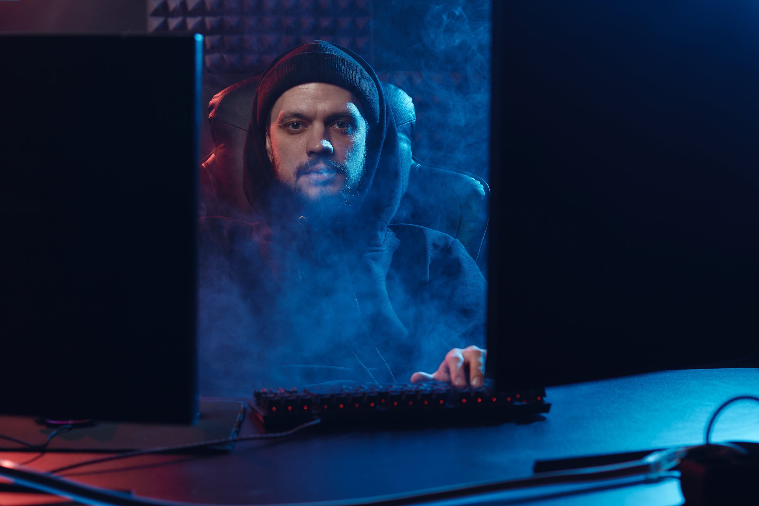 Mężczyzna w czapce siedzi przed komputerem w zaciemnionym pokoju, oświetlenie podkreśla dym unoszący się wokół niego.