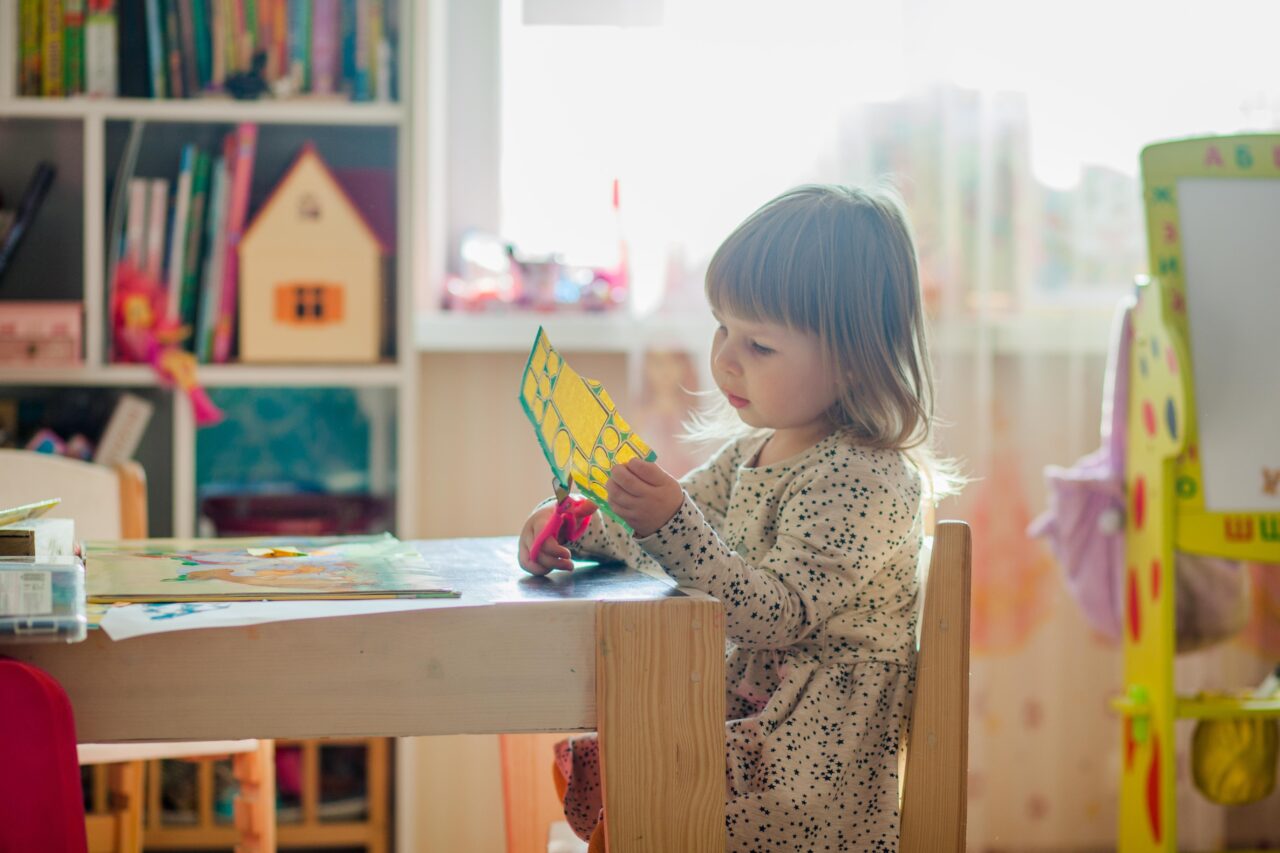Mała dziewczynka siedzi przy drewnianym stole i trzyma w rękach kolorową papierową kreaturę, w tle widoczna jest rozmyta półka z książkami i zabawkami w jasnym pokoju zabaw.