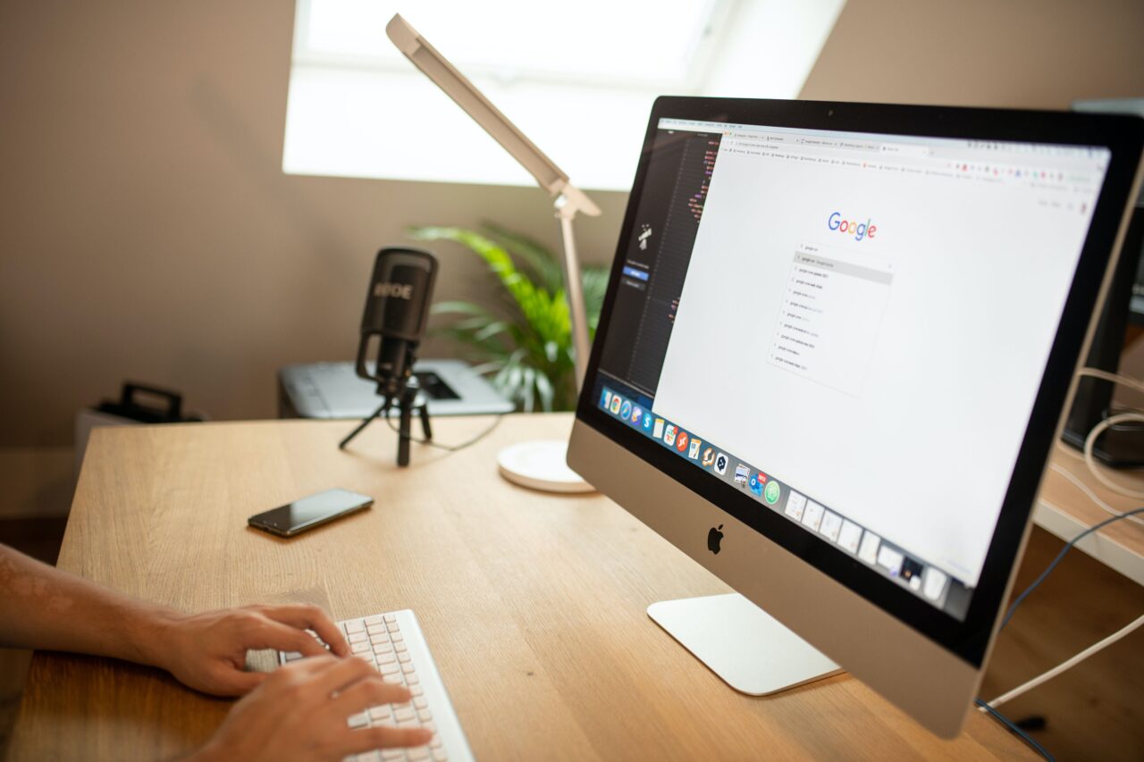 Biurko z komputerem iMac, na którym wyświetlana jest strona główna Google, obok klawiatura i smartfon, w tle mikrofon pojemnościowy i biurowa lampa.