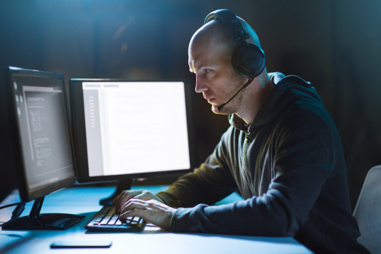 Mężczyzna programista pracuje przy komputerze w ciemnym pomieszczeniu, nosząc słuchawki na uszach. Sprawdza jak wykryć podsłuch na telefonie