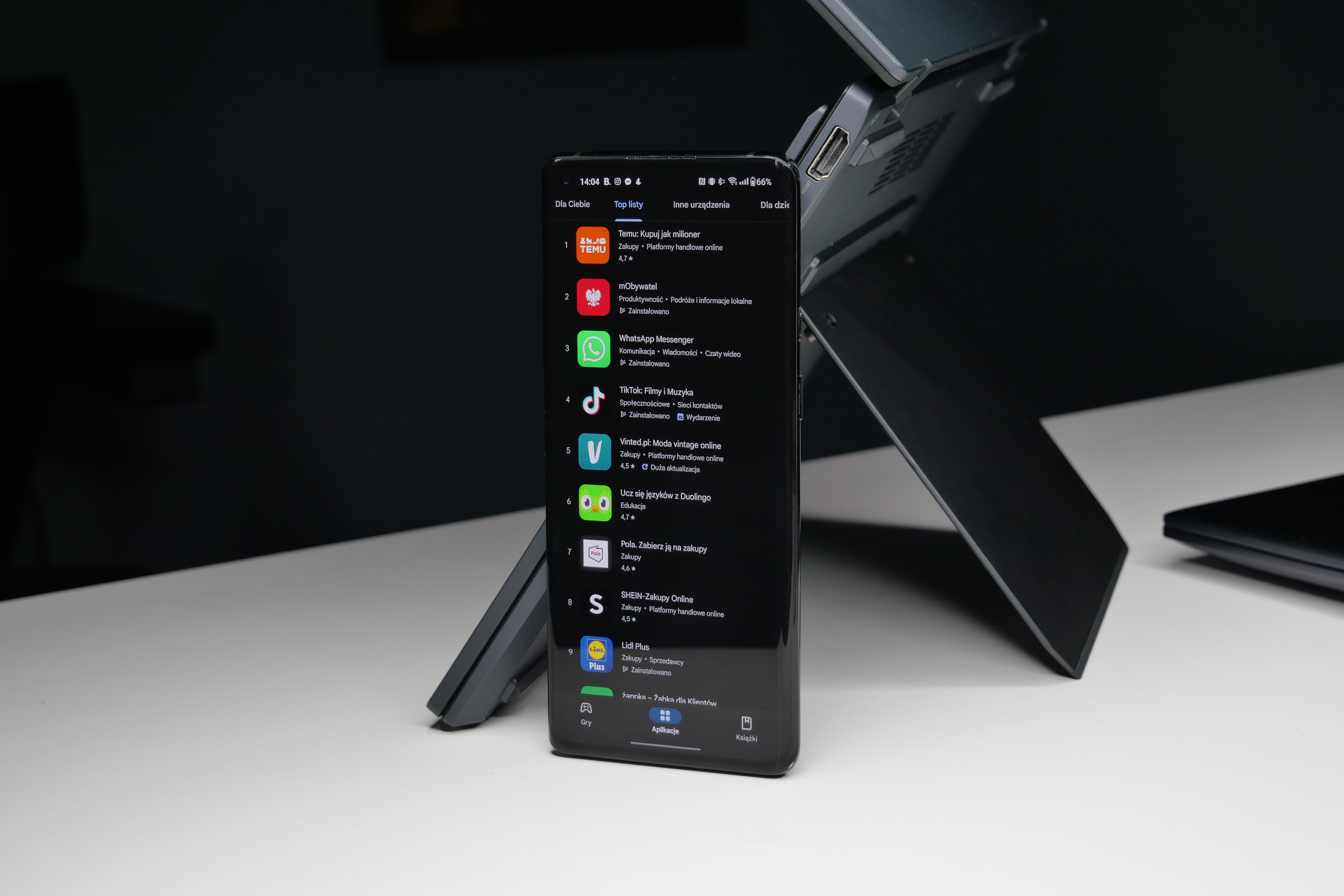 Czarny smartfon stoi na składanym stojaku, wyświetlając ekran z aplikacjami na tle biurka.