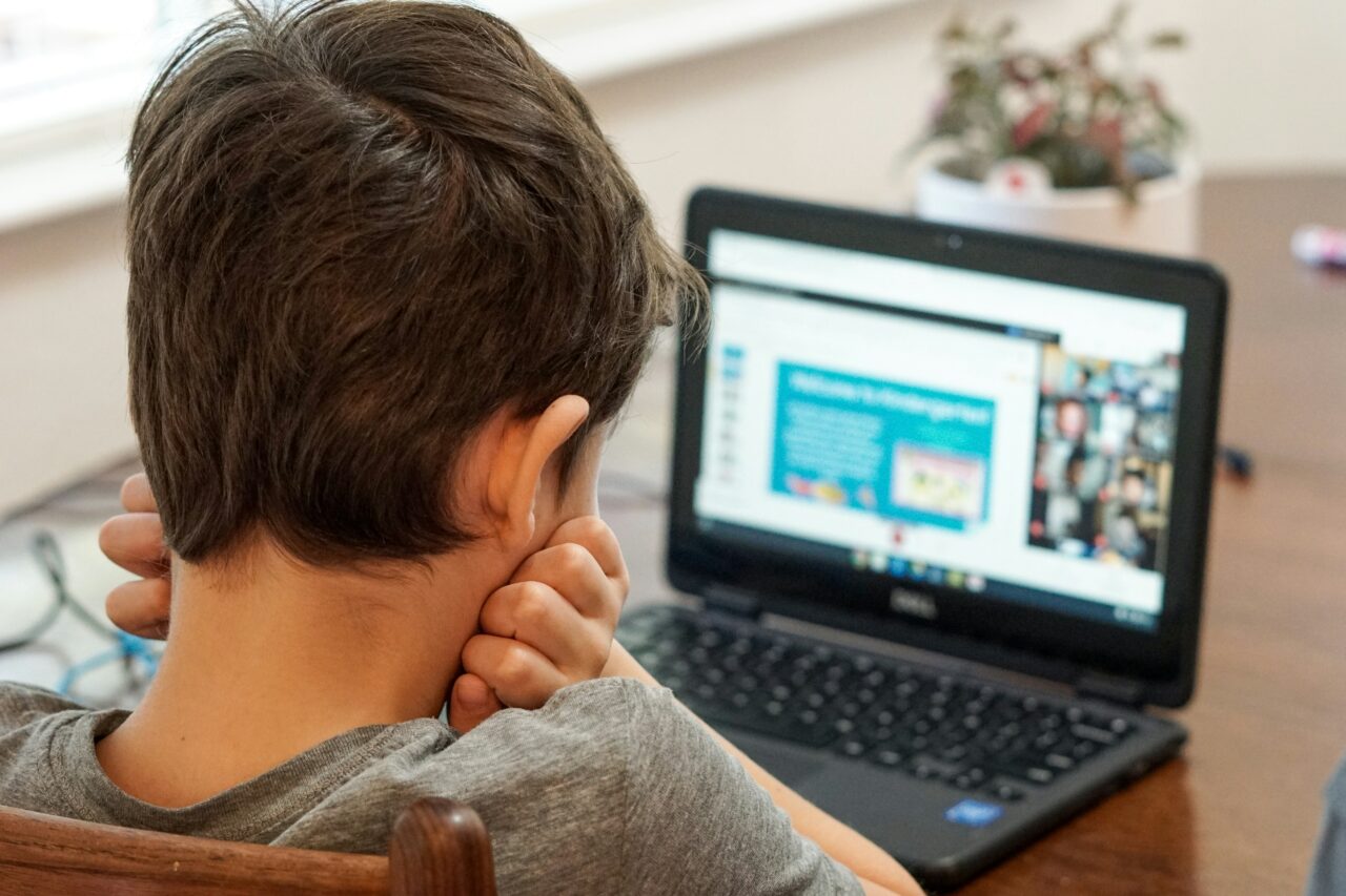 Dziecko siedzi przed laptopem i uczestniczy w wideo lekcji, wspierając głowę ręką.