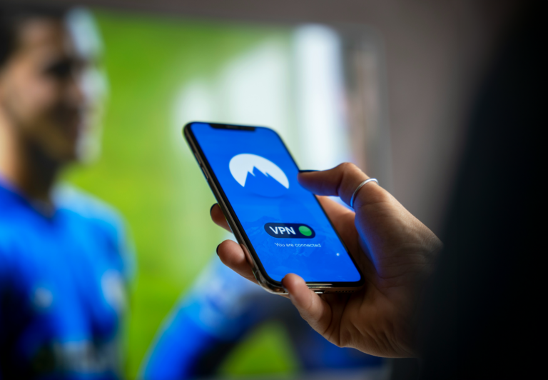 Osoba trzymająca smartfon z ekranem pokazującym aktywną aplikację VPN, w tle nieostro sportowiec w niebieskim stroju.