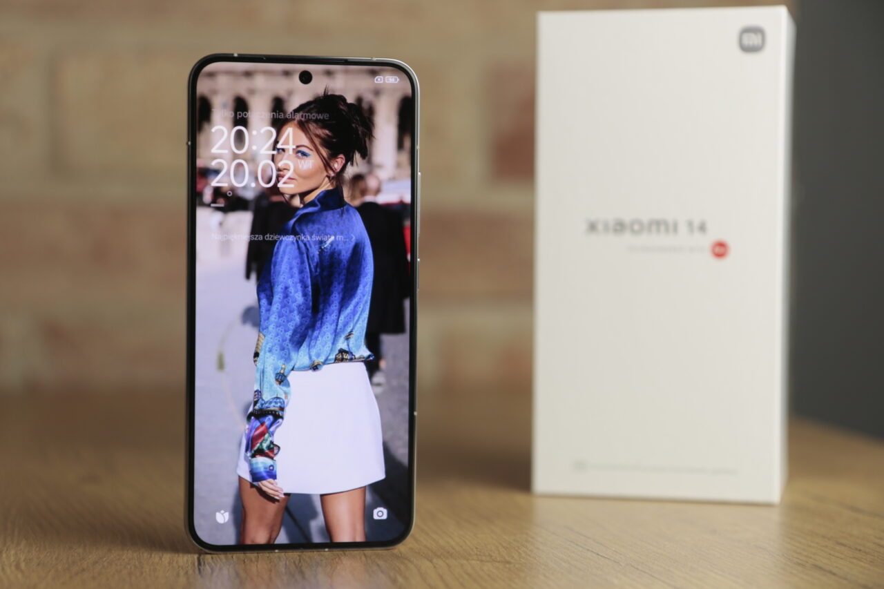Smartfon Xiaomi 14 na pierwszym planie z wyświetlonym obrazem kobiety, w tle rozmyte opakowanie produktu.