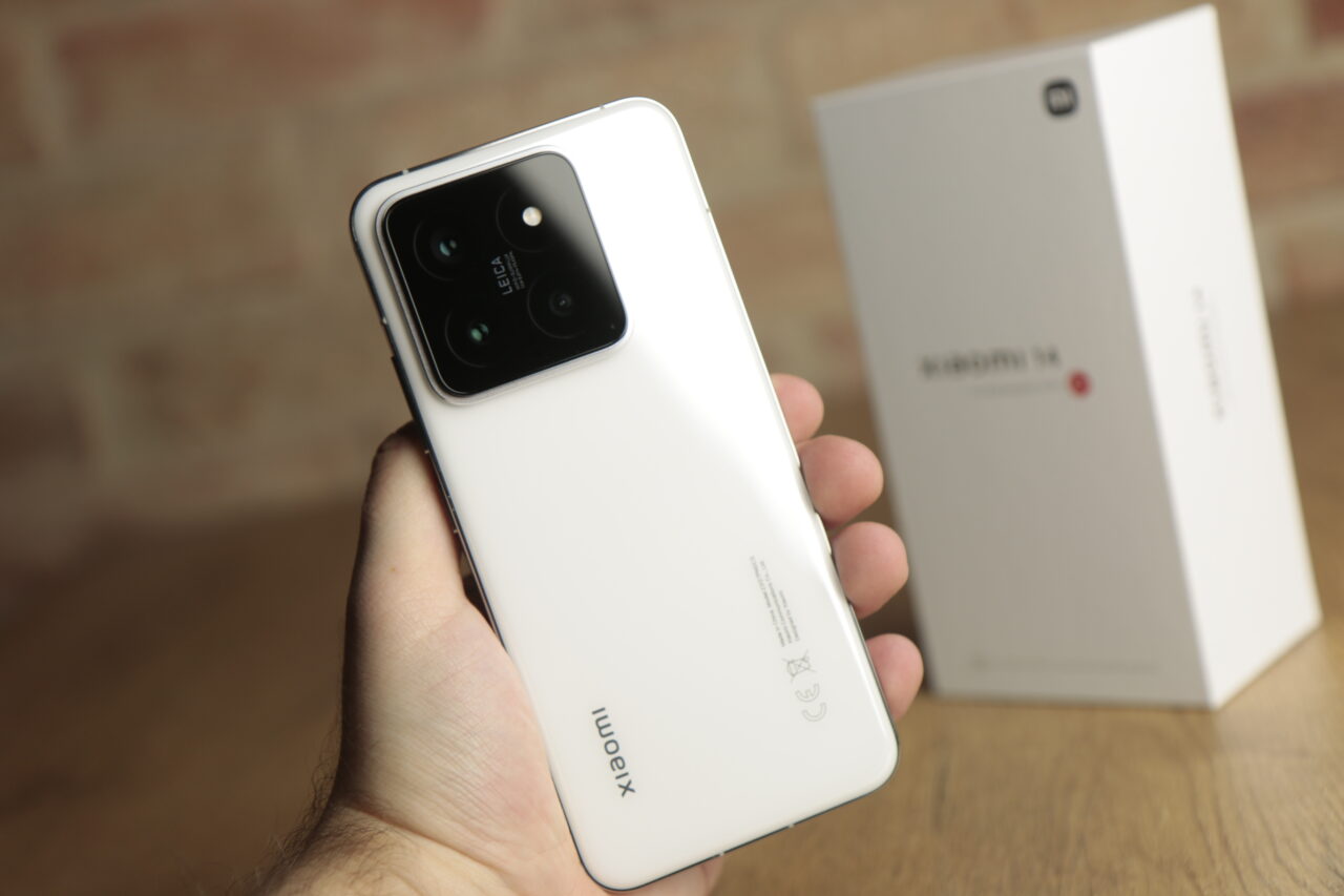 Biała tylna część smartfona z poczwórnym aparatem marki Leica trzymanego w dłoni, w tle rozmyte opakowanie produktu.