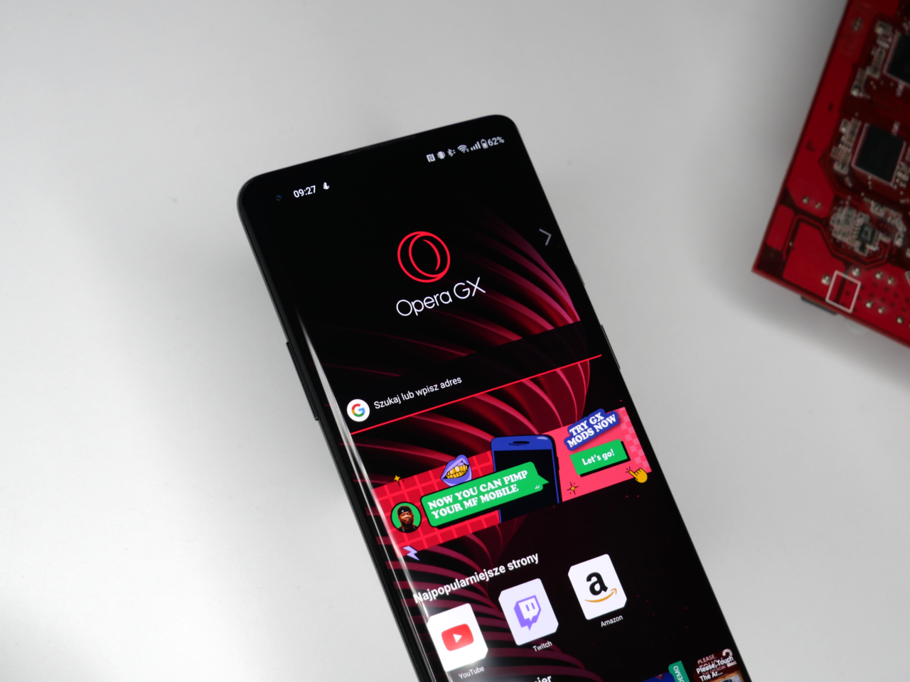 Smartfon leżący na białym tle z otwartą aplikacją przeglądarki Opera GX na ekranie, obok częściowo widocznych elektronicznych komponentów na czerwonym pcb.