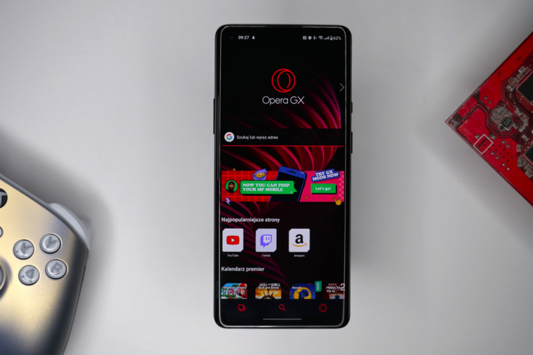 Smartfon leżący na białym stole z otwartą aplikacją przeglądarki Opera GX, obok fragment kontrolera do gier oraz czerwona płyta elektroniczna.