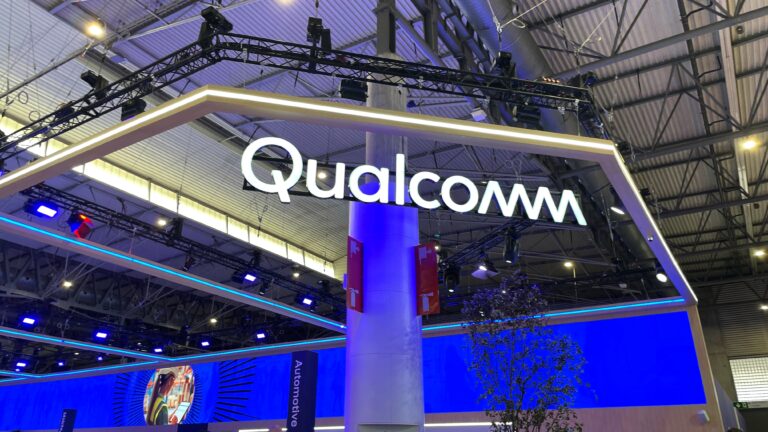 Stoisko firmy Qualcomm na targach z dużym, podświetlanym logo firmy na górze, otoczone oświetleniem scenicznym i niebieskim tłem.