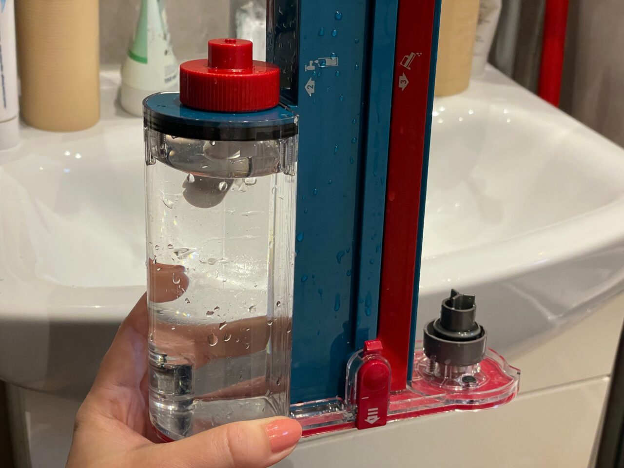 Ręka trzymająca przezroczysty pojemnik z wodą obok niebieskiego i czerwonego poziomica na białej umywalce.