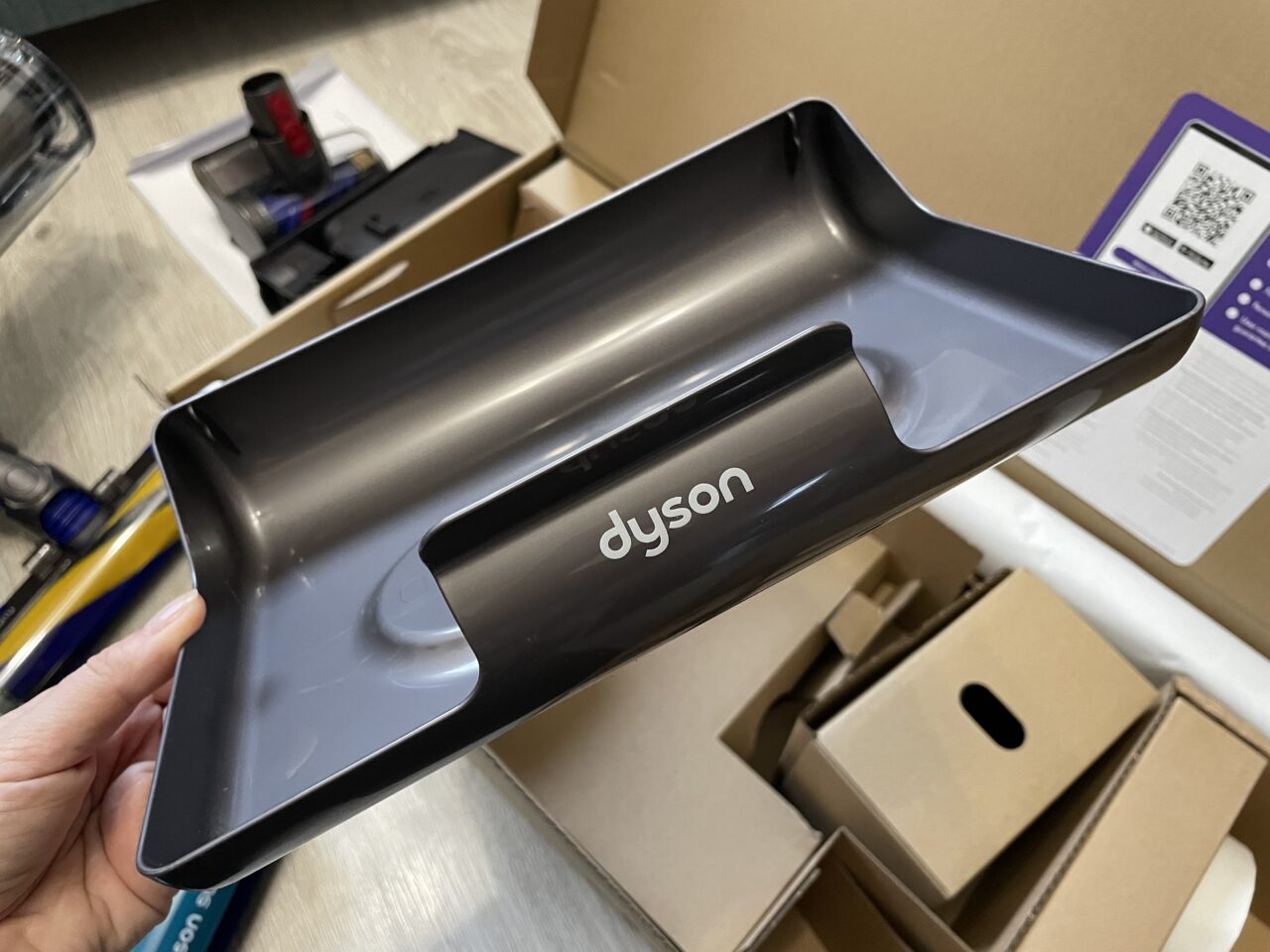 Osoba trzyma szary pojemnik do odkurzacza marki Dyson nad otwartym pudełkiem z różnymi akcesoriami do sprzątania.