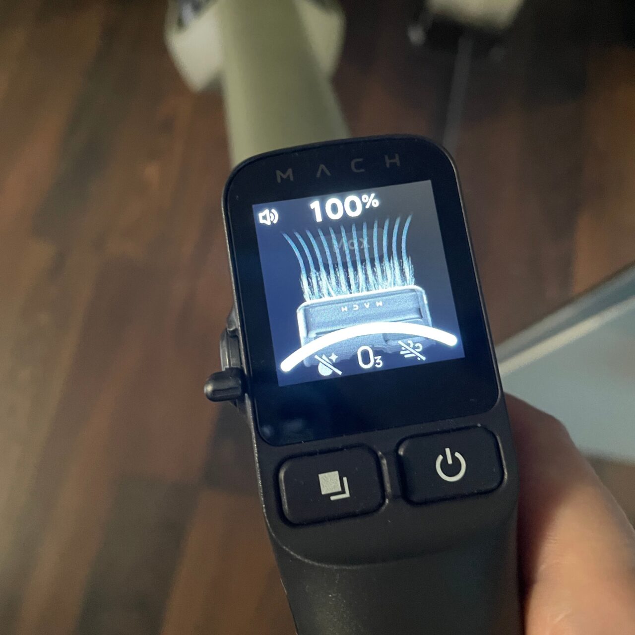 Czyjś dłoń trzymający maszynkę do golenia z wyświetlaczem pokazującym naładowanie baterii w 100% oraz ikonę ostrza.