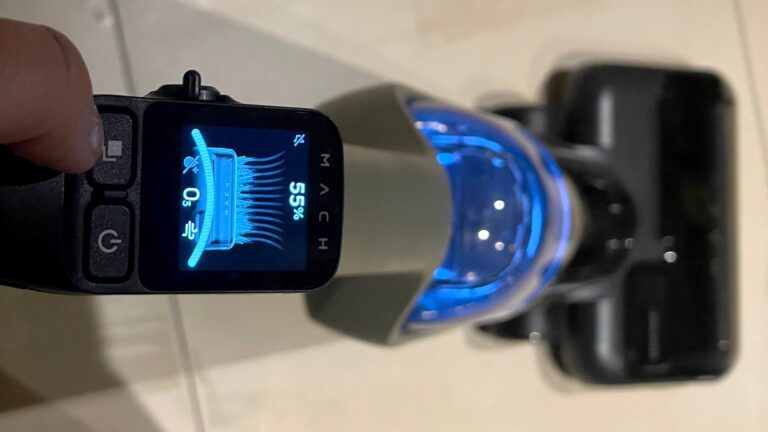Pierwszoplanowy widok na wyświetlacz elektroniczny ręcznego urządzenia, pokazujący niebieskie światło i procent naładowania baterii (55%), trzymanego przez rękę z rozmytym tłem.