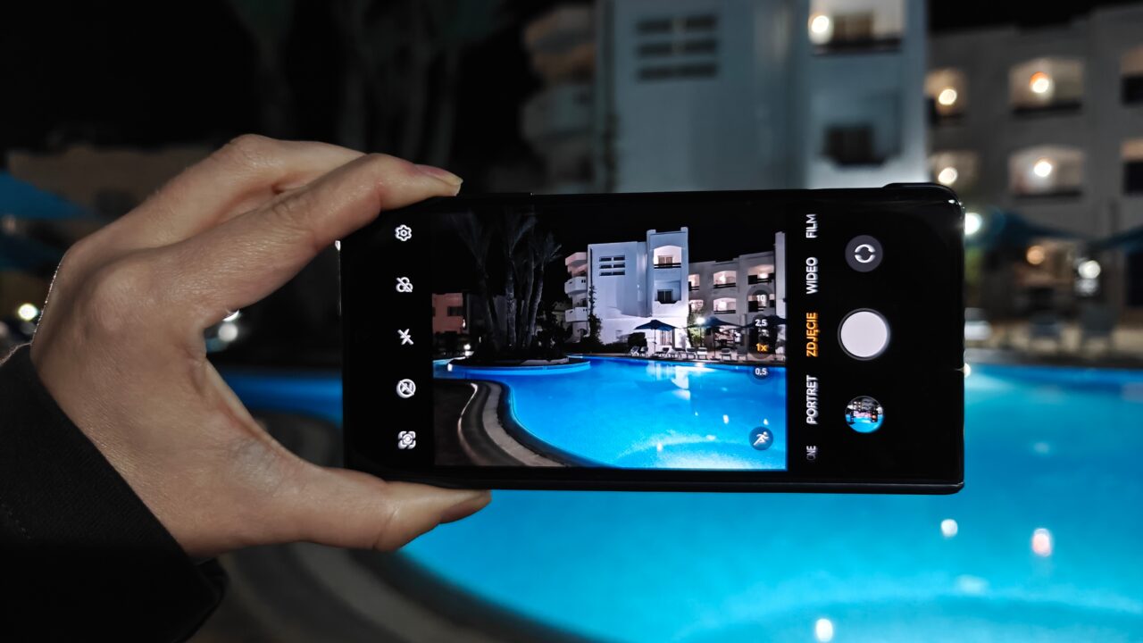 Dłoń trzymająca smartfon, na którego ekranie wyświetlany jest widok basenu oświetlonego nocą z budynkami w tle.