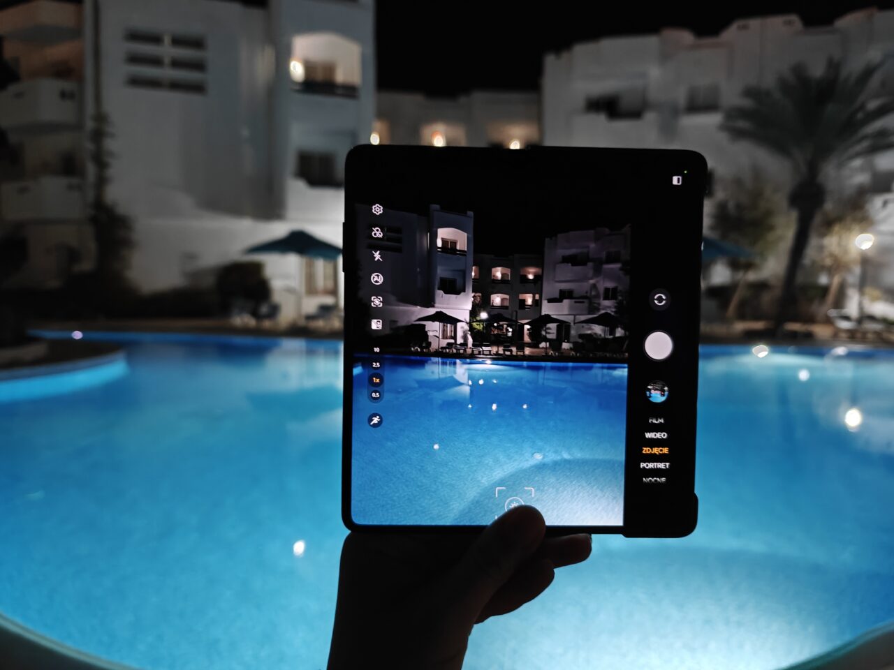 Osoba trzyma tablet z widokiem na basen i białe budynki nocą, obraz z tabletu jest wyświetlany w trybie kamery.