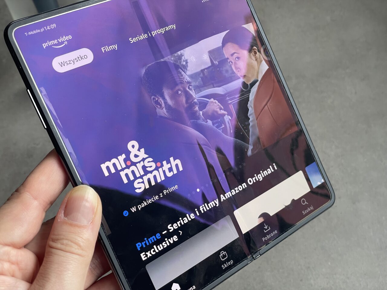 Dłoń trzymająca smartfon z otwartą aplikacją Prime Video wyświetlającą miniaturę filmu "Mr. & Mrs. Smith" z opcją "W pakiecie z Prime" oraz oznaczeniem "Prime – Seriale i filmy Amazon Originals".