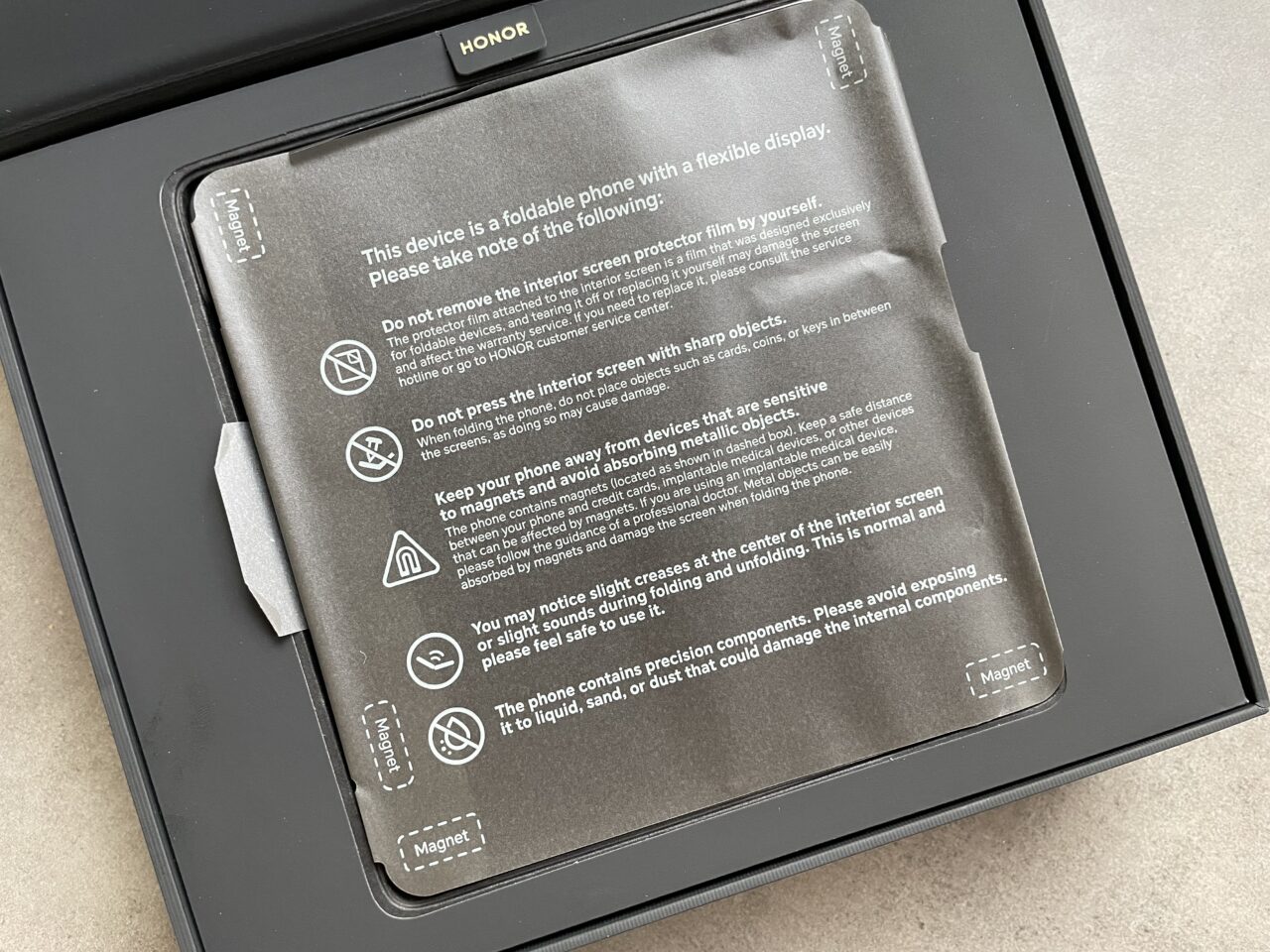 Skladany telefon Honor umieszczony w opakowaniu z plastikowym etui ochronnym, na którym znajdują się ostrzeżenia i wskazówki dotyczące użytkowania, w tym unikania kontaktu z cieczą, magnesami i ostrymi przedmiotami.