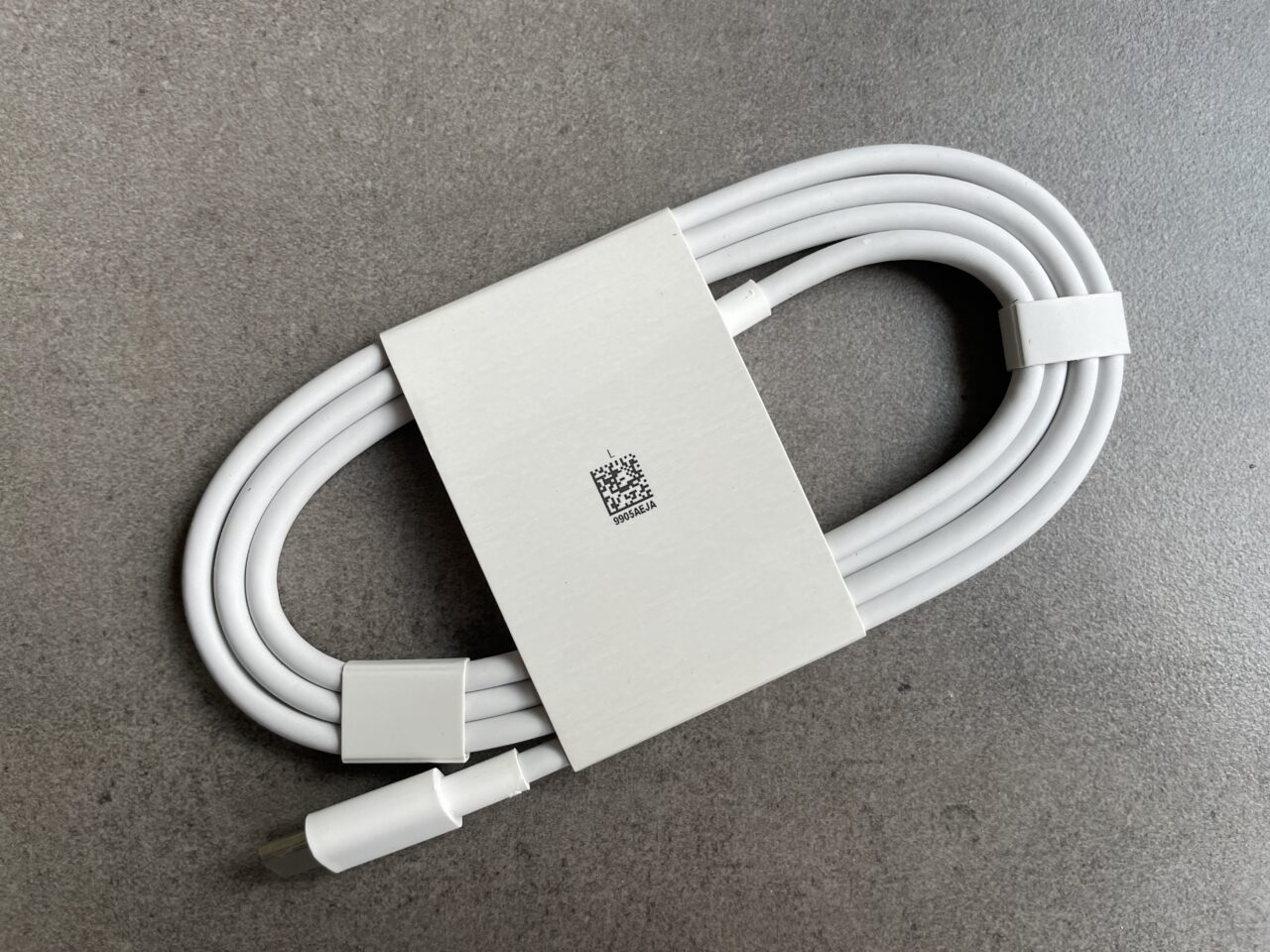 Zdjęcie białego, zwiniętego kabla zasilającego z wtyczką USB typu C i prostokątnym adapterem z kodem QR, leżące na szarym tle.