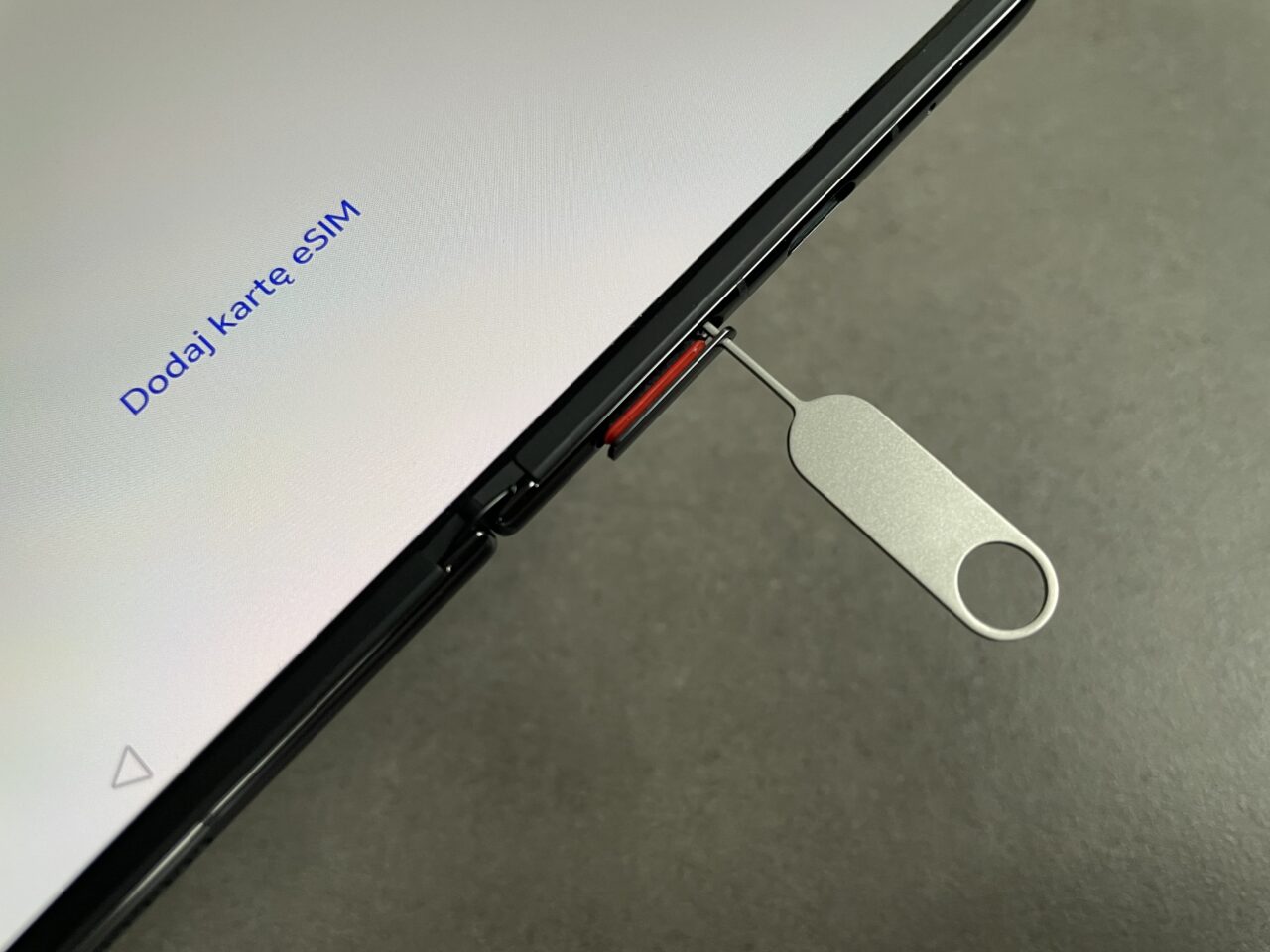 Zdjęcie ekranu telefonu z napisem "Dodaj Kartę eSIM" i fragmentem wyciąganego tacki na kartę SIM z użyciem metalowego narzędzia.