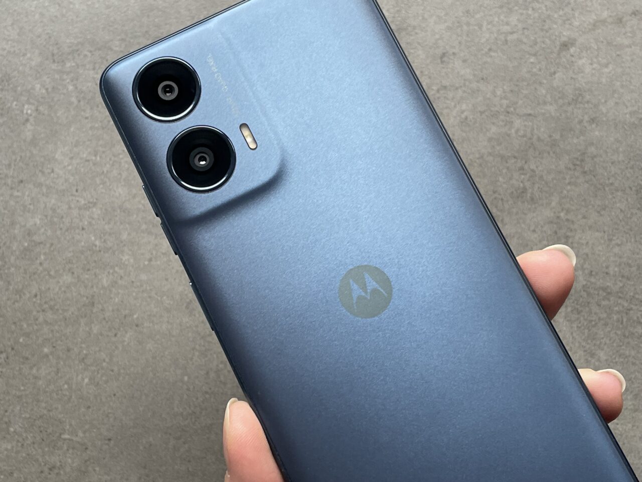 Tylna część smartfona marki Motorola w kolorze niebieskim, z podwójnym aparatem i logo producenta, trzymana w dłoni.