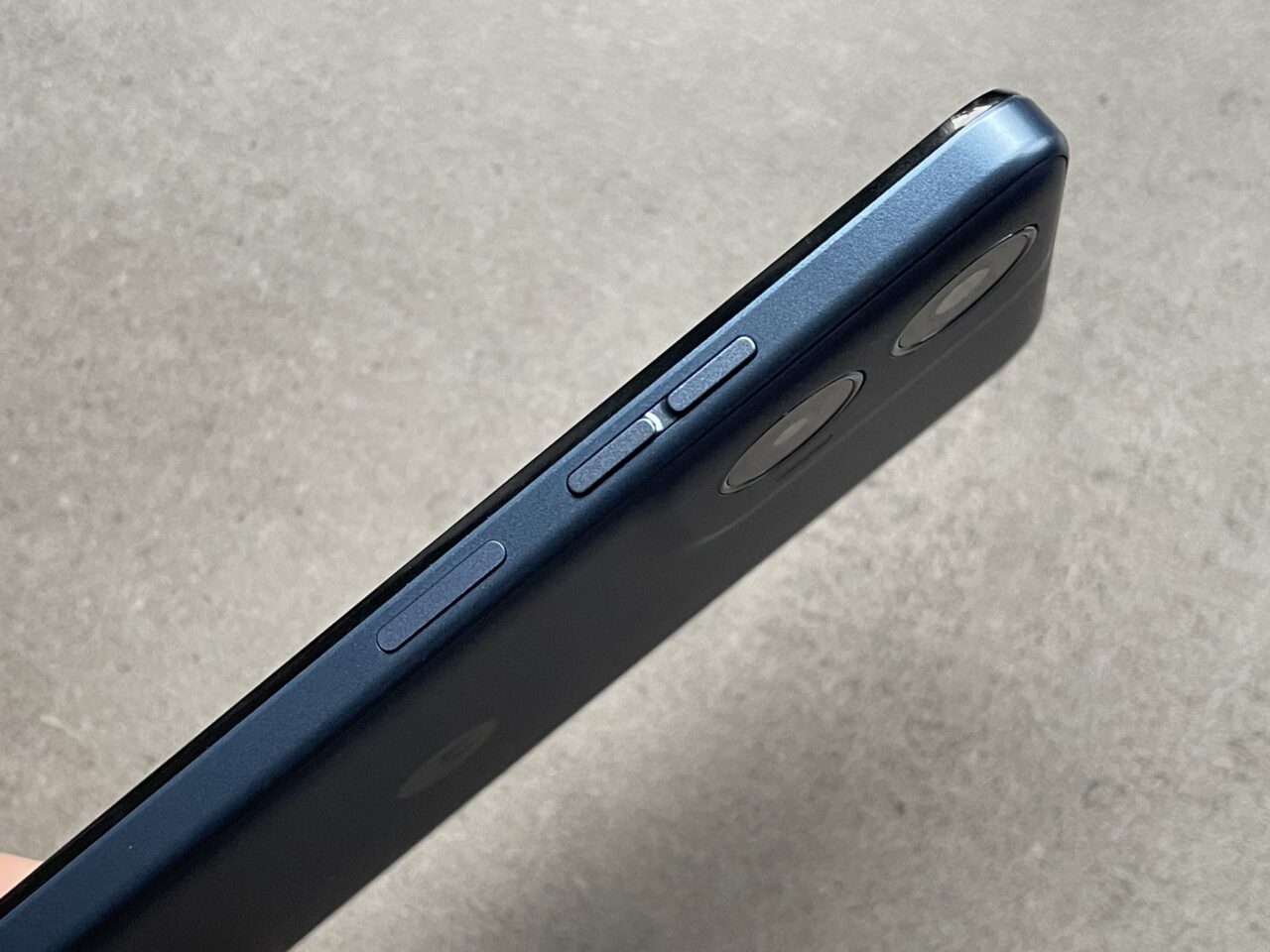 Ciemnoniebieski smartfon trzymany bokiem, ukazujący przyciski głośności i przełącznik trybu cichego oraz zaokrąglony narożnik z kamerami.